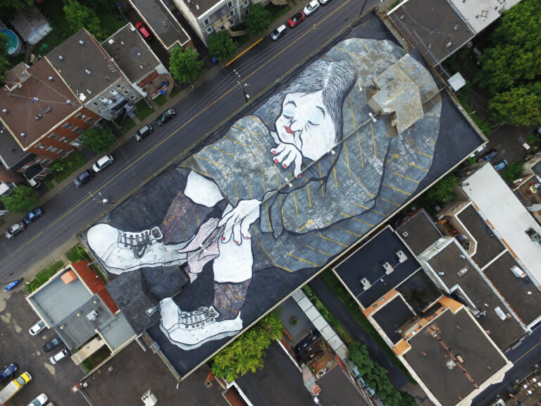 Une fresque murale géante d'Ella et Pitr représentant un personnage allongé sur un toit d'immeuble, vue aérienne.